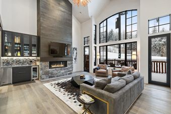 A modern and spacious living room with a large stone fireplace, a wet bar, comfortable seating, and floor-to-ceiling windows showcasing a snowy forest.
