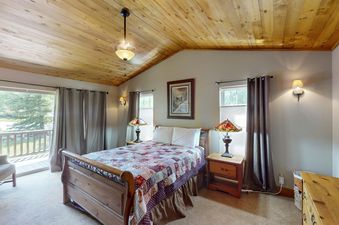 The cozy master bedroom at Bear Creek Lodge Townhome features a queen bed with a patchwork quilt, a large window with views of the surrounding trees, and a side table with a lamp.