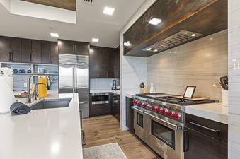 A modern kitchen with dark wood cabinets, stainless steel appliances, a Wolf range with red knobs, white countertops, and a veined stone backsplash.