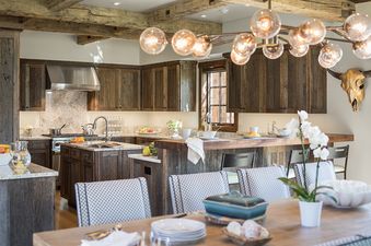 A rustic-modern kitchen with wooden cabinetry, granite countertops, and a large island, leading to a dining area with an elaborate chandelier.