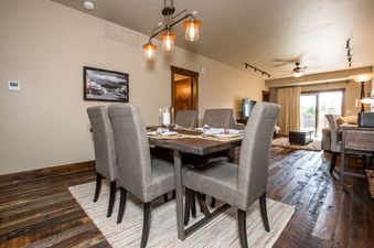 The dining area in Baker Loft 203 features a wooden table with six gray upholstered chairs, an industrial-style chandelier, and a large sliding glass door leading to an outdoor view.