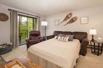 The living area of Beaver Village 823 features a comfortable brown sofa that converts into a bed, a brown recliner, and direct access to a balcony with lush green views.