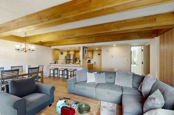 A spacious open-concept living area with a gray sectional sofa, a matching armchair, a dining table with chandelier, and a kitchen with a breakfast bar.