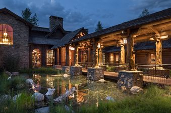 The Royal Wulff Lodge at dusk, showcasing its stone and timber architecture, illuminated walkways over a reflective pond, and surrounding natural landscape.