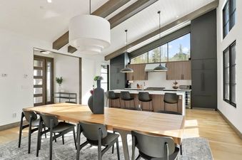 A modern open-plan dining room and kitchen with a large wooden dining table, black chairs, and a kitchen island with bar stools, all under a vaulted ceiling with exposed beams and large windows showcasing an outdoor view.