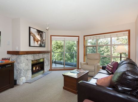 A spacious and inviting living room at Aspens on Blackcomb Unit 415, featuring a cozy fireplace, comfortable brown leather sofa, and large windows with lush green outdoor views.