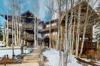 The exterior of Settlers Lodge, a ski-in ski-out condo building, features a stone and wood exterior with snow-covered grounds and a walkway leading up to the front entrance.