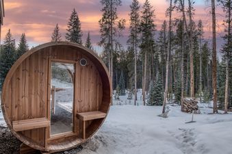 A barrel sauna with a glass door stands in a snow-covered forest with tall pine trees under a pink and purple sunset sky.