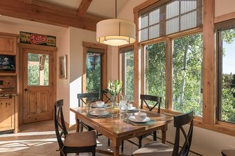 A cozy dining area with a wooden table set for four, surrounded by large windows offering views of green trees and a distant ski lift.