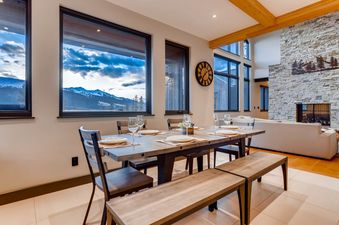 A spacious dining area with a long wooden table and benches, set with plates and glasses, offering views of snowy mountains through large windows.