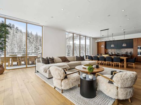 The spacious great room at Founders Place 1701 features a cozy living area with a sectional sofa and fur-upholstered chairs, alongside a dining area and kitchen, all framed by large windows revealing a snowy mountain landscape.