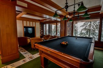 A pool table with racked balls stands ready in The Totem Chalet's games room, offering a view of snowy pine trees through large windows.