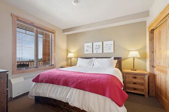 The king bedroom in Morning Eagle 207 features a bed with white linens, a pink throw blanket, two nightstands with lamps, and a window with mountain views.