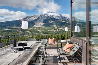 An expansive outdoor balcony, featuring a rustic wooden dining table, modern chairs, and stylish lamps, offers breathtaking panoramic views of snow-capped mountains and a dense forest under a clear sky.