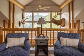 A cozy living area features two blue armchairs, a wooden side table with a lamp, and taxidermy deer heads, all set against a backdrop of large windows with mountain views.