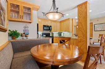 A cozy dining nook with a round wooden table and a built-in upholstered bench is situated next to a kitchen with light wood cabinets and black appliances in The Charter G140.