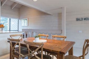 A wooden dining table with six chairs sits in a dining room with white walls and a wooden sign that reads "Bald Mountain".