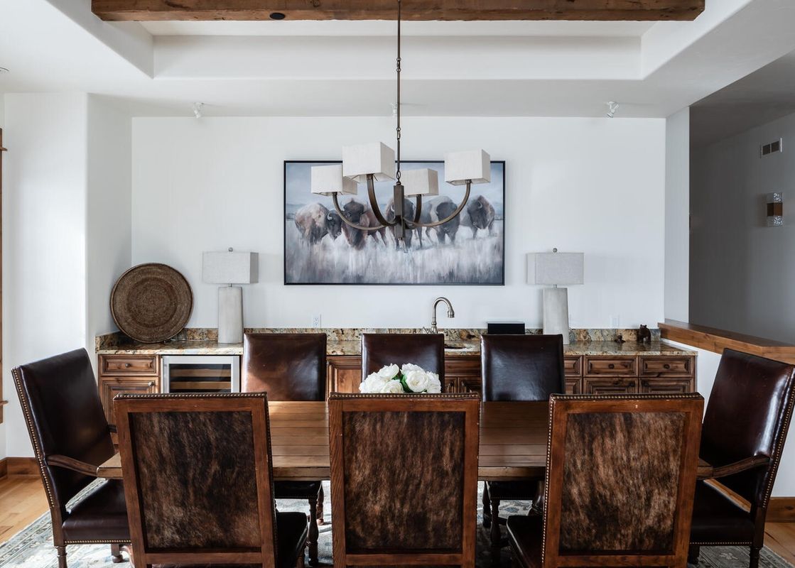 The dining table of Moonlight Basin Luxury Suite 6A is set with brown leather chairs, and there are wooden cabinets and a wine fridge in the background.
