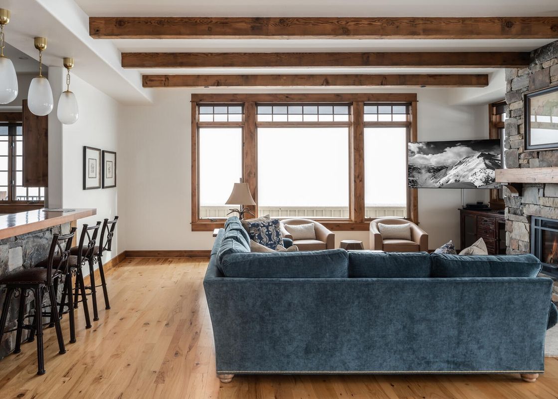 The living room features a large, comfortable sofa, a stone fireplace, and windows with views of the snowy mountain peaks.