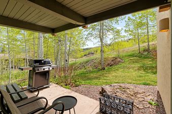 A private patio at Top of the Village Leaf 401 with an outdoor grill, two chairs, and a small table, surrounded by aspen trees and a green hillside.