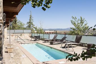 The outdoor pool and hot tub area at Lowell 402A, featuring a sparkling pool, an inviting hot tub, comfortable lounge chairs, and a patio table, all set amidst lush trees with distant mountain views.