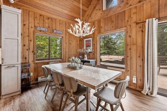 A rustic yet elegant dining area in The Village House with a large wooden table, antler chandelier, and forest views from expansive windows.