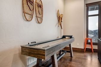 A shuffleboard table with shuffle pucks and accessories is set up in a room with light-colored walls and dark wood floors, featuring snowshoes as decor and a window offering a snowy mountain view.