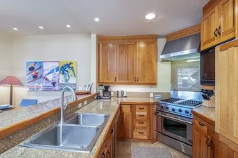 A modern kitchen with granite countertops, light wood cabinetry, and stainless steel appliances is shown, featuring a double sink and a gas range.