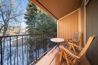 The balcony at Beaver Village 332 features a table and two chairs, all lightly dusted with snow, overlooking a snowy, treed landscape.