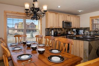 A large, wooden dining table set with plates and glasses sits in front of a window, offering a view of houses and trees outside, while a kitchen with wooden cabinets and stainless steel appliances is visible in the background.