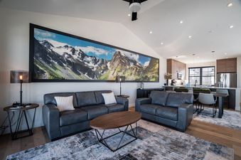A spacious living room at Penthouse in the Pines, featuring comfortable sofas, a large mountain landscape artwork, and an open-concept kitchen with modern appliances and bar seating.
