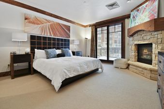 A large king-size bed with a dark wooden headboard and white bedding is centered in a spacious bedroom, with a stone fireplace to the right and a balcony visible through large glass doors.