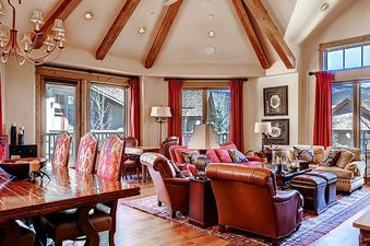 The Arrowhead Village Condominium Platinum 5 Bedroom shows a spacious living area with hardwood floors, a large wooden dining table, comfortable sofas, and large windows with red curtains offering views of a snowy landscape.