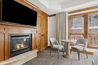 A cozy living area in The Aspen 44A featuring a fireplace with a TV above, a small dining table with two chairs, and large windows looking out onto a snowy exterior.