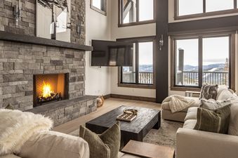 The living area at 22 Lower Cascade Ridge features a stone fireplace, a large flat-screen television, and expansive windows that offer stunning views of the snow-covered mountains.
