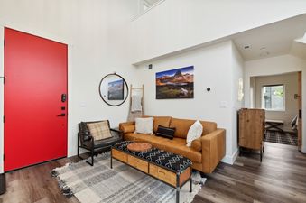 A modern and bright living room in Quarry A25 with a red entrance door, a tan leather sofa, and a mountain landscape artwork.