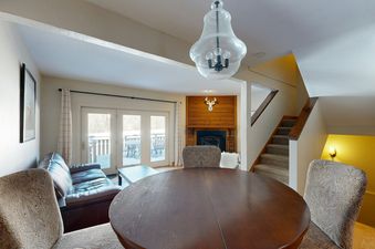 The dining room at The Fairway Serenity features a large wooden table and chairs, and a view into the living room with a fireplace and a couch.