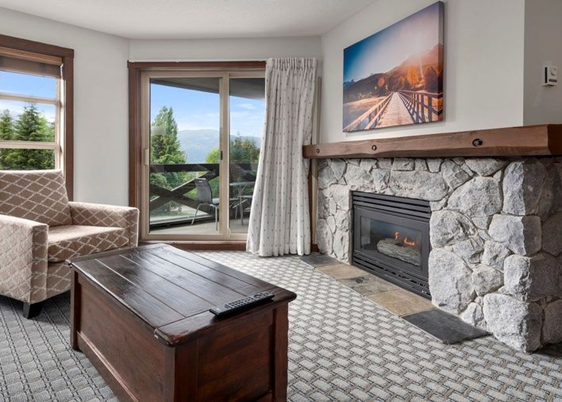 A cozy living room at Blackcomb Springs Two Bedroom Slopeside Suite features a stone fireplace, a comfortable armchair, and a dark wood coffee table, with a large window and glass door offering mountain and forest views.