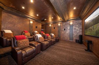 A home cinema with plush brown leather recliners, each adorned with a red or patterned pillow and a white blanket, is illuminated by warm ceiling lights, showcasing a large screen displaying a mountain landscape.