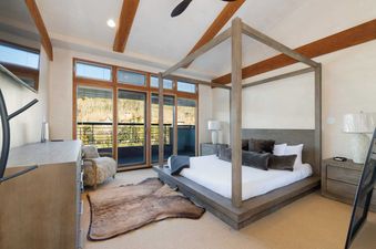A master bedroom at Peaks Penthouse 617 features a large, modern four-poster bed with a view of mountains and trees through a large sliding glass door to a balcony.