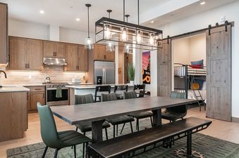 A modern kitchen and dining area with a long table and chairs, leading into a bunk room behind barn doors.
