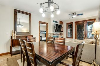 The image shows the dining area of BlueSky Breckenridge 407, featuring a wooden dining table and chairs, with a living area and fireplace in the background.