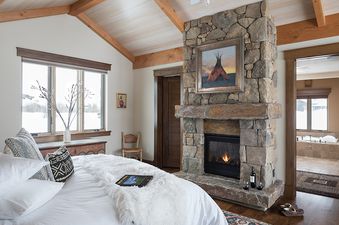 A cozy bedroom at Paintbrush Retreat features a stone fireplace, a large bed with white linens, and a window overlooking a snowy landscape.