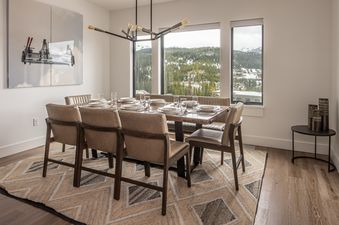 A modern dining room with a long wooden table set for eight, featuring large windows with views of snow-covered mountains and pine trees.