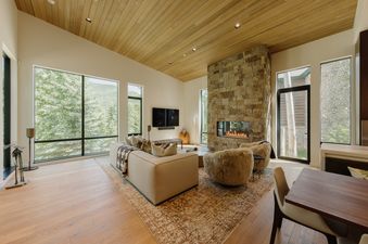 The living area of Vail Aspen Haven features a comfortable sofa and two fur-covered chairs in front of a stone fireplace, with large windows offering views of the lush green trees outside.