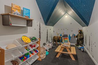 A play area for children with a small wooden table and chairs, stuffed animals, and a variety of toys in a bin.  The ceiling is painted blue and has white stars on it. The walls are painted white and have a woodland mural on them.