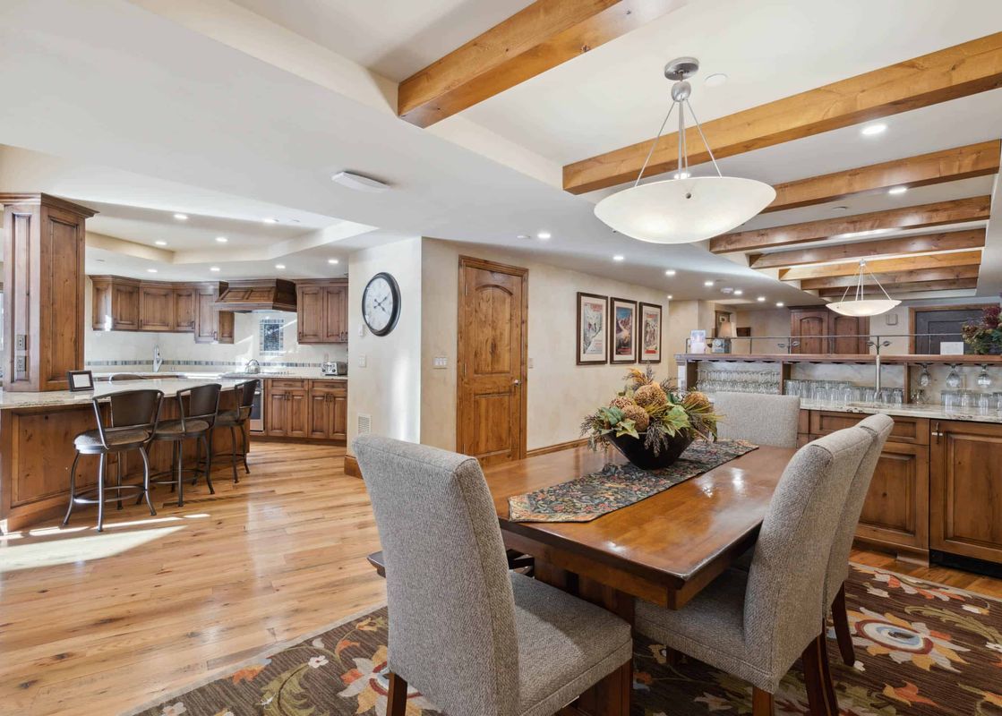 A spacious dining area with a long wooden table and upholstered chairs, leading into a modern kitchen with a large island and bar stools, all under a ceiling with exposed wooden beams.