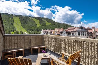 A rooftop patio at Vail 21 601 with wooden furniture overlooking a mountain with ski slopes and village buildings under a blue sky.