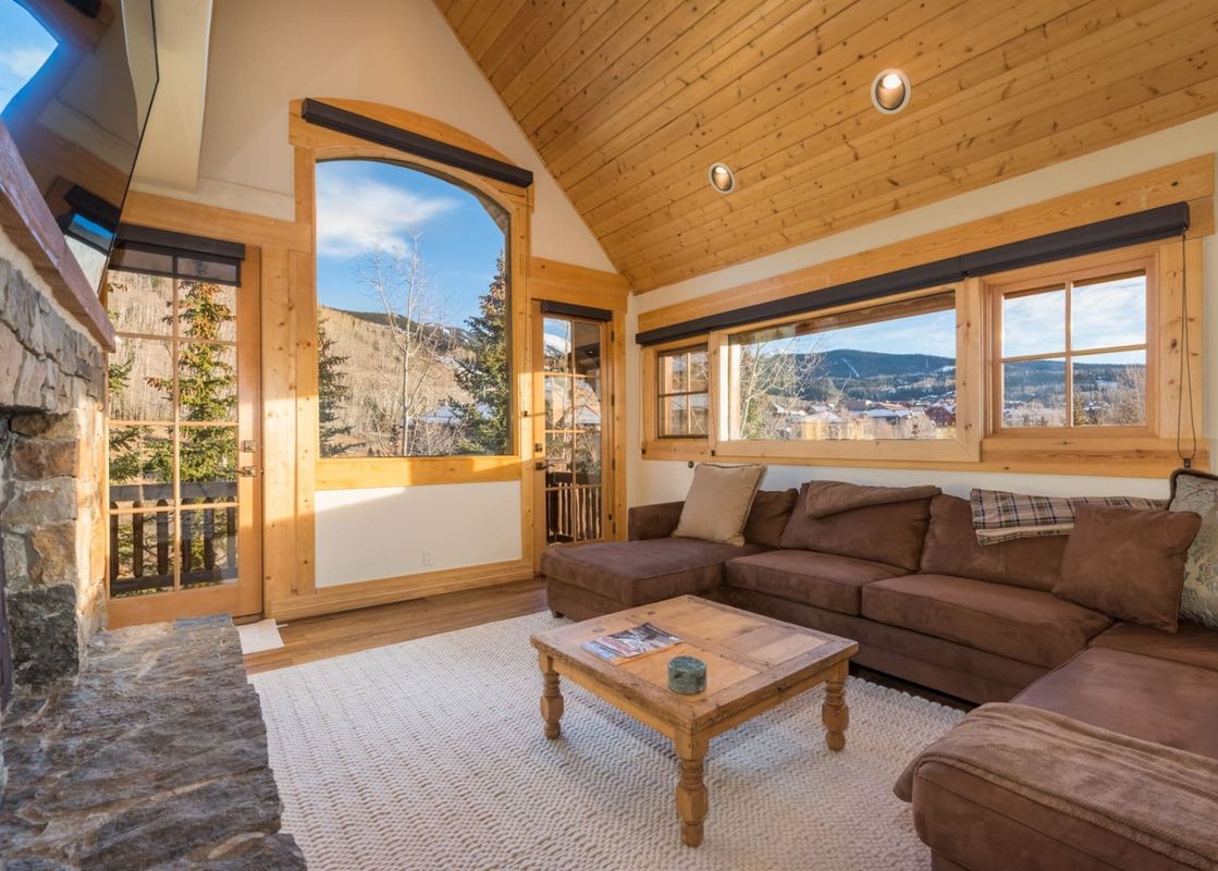 A cozy living room in See Forever Residence 110 with a large brown sectional sofa, a wooden coffee table, and expansive windows offering mountain views.