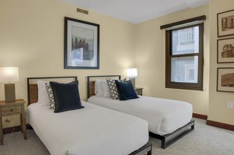 A twin bedroom at Lowell 203 featuring two beds with white linens and navy pillows, a bedside table with a lamp, and framed artwork on light beige walls.